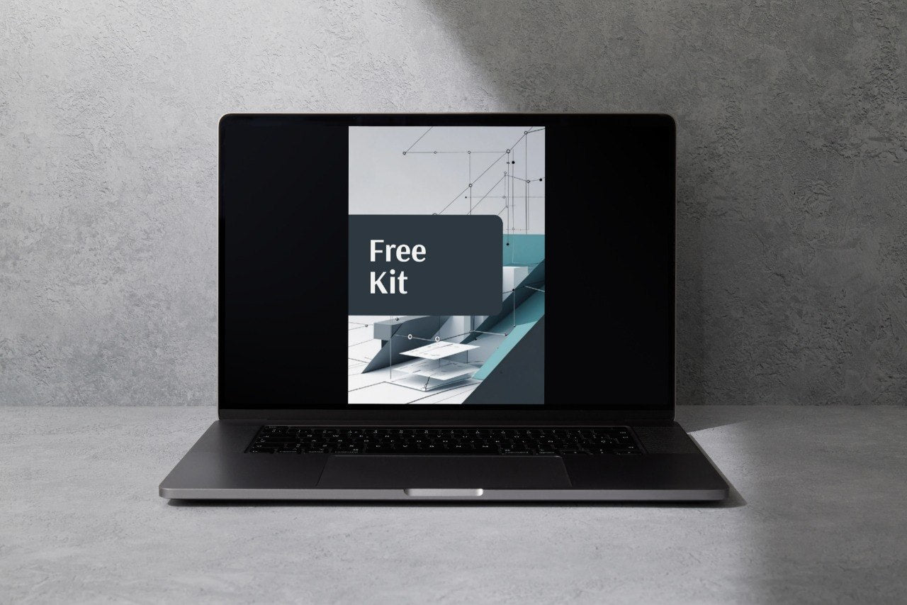 An open black laptop sits on a gray concrete surface against a textured gray wall, with a screen graphic reading 'Free Kit' displayed on the screen.