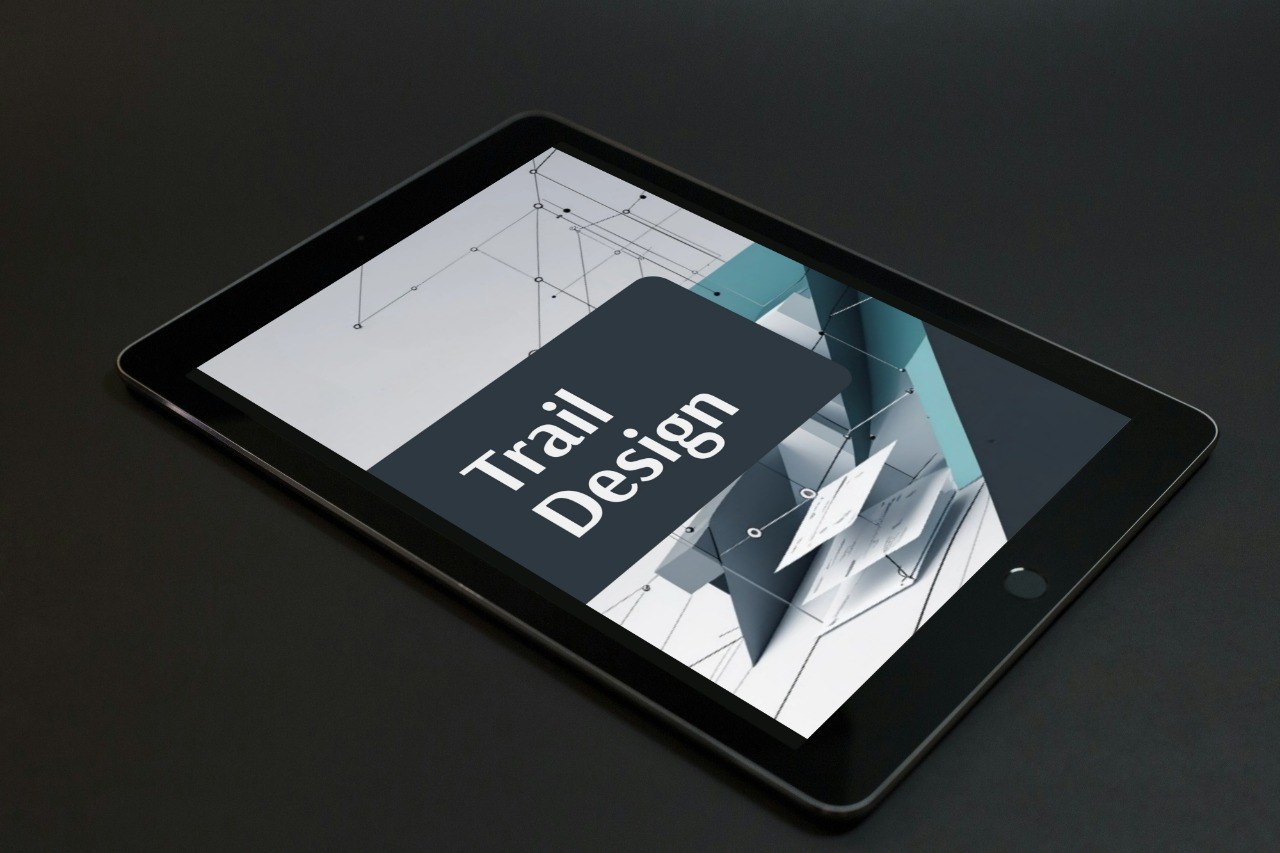 A black tablet device shown at an angle on a dark surface, displaying a digital cover design with the words 'Trail Design' over a geometric abstract graphic.